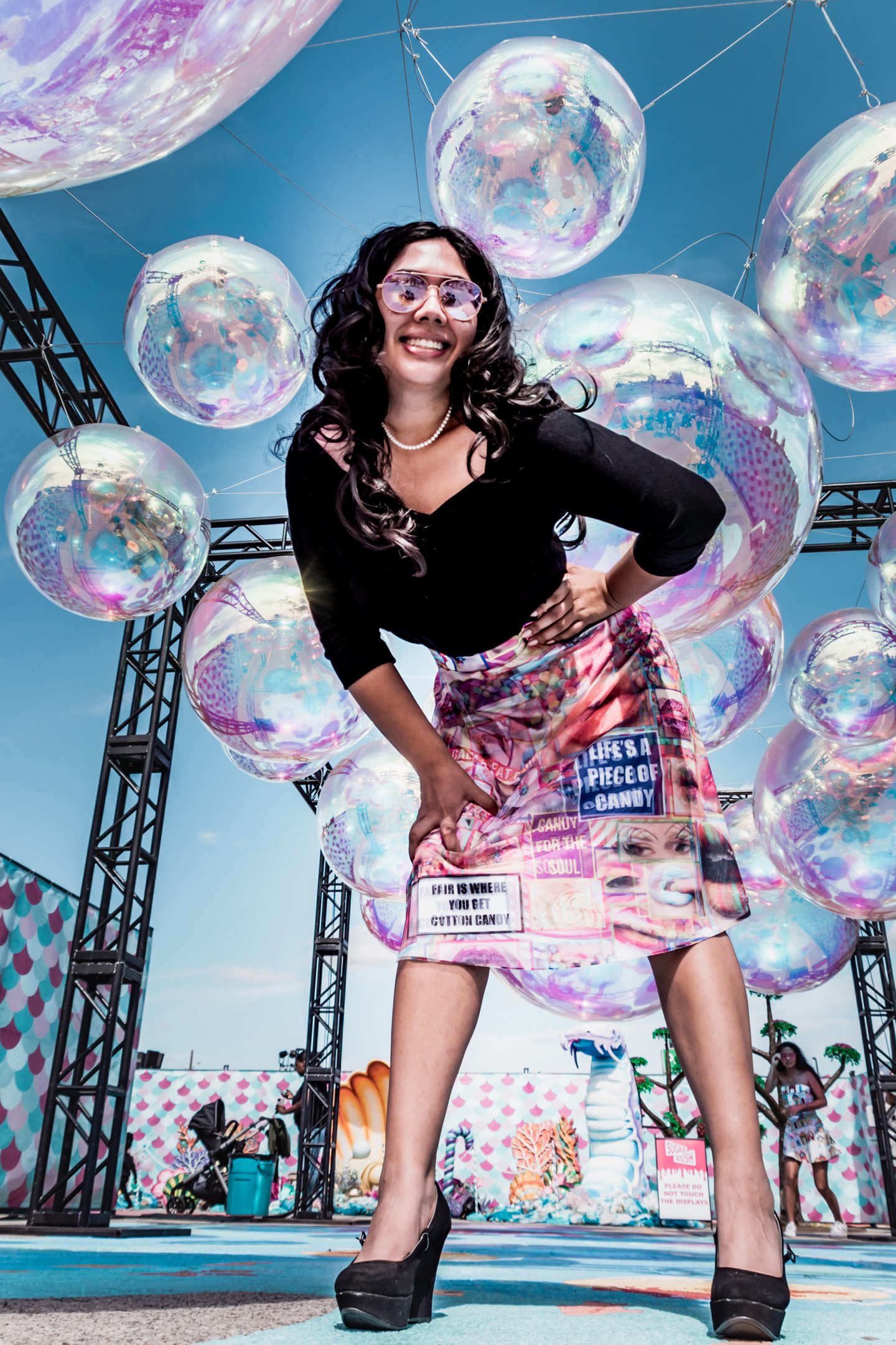 Portrait of woman in a holographic skirt and black shirt, leaning forward into view. Giant fake bubbles are held in the air behind her.