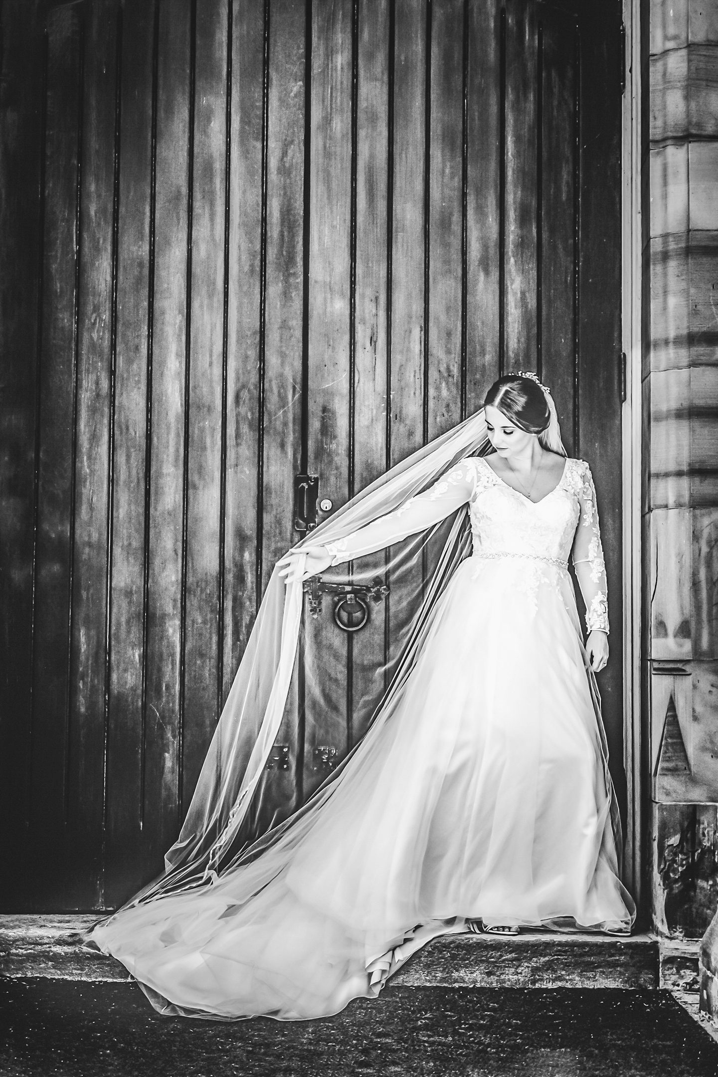 Bride standing in a wooden doorway looking down at her hand as it plays with her veil. Black and White image